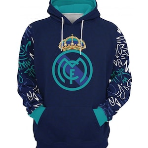 Op de afbeelding: Een marineblauwe hoodie met een teal groene capuchon en bies. De hoodie heeft een witte afbeelding van het Real Madrid-wapen met een kroon erop. De mouwen hebben een wit grafisch patroon.