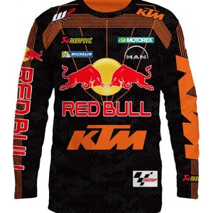 Puede incluir: Camiseta negra de manga larga con detalles en naranja y blanco. La camiseta presenta el logotipo de Red Bull, el logotipo de KTM y otros patrocinadores como Akrapovic, Michelin y Motorex. La camiseta también tiene el texto "Red Bull" y "KTM" en ella.