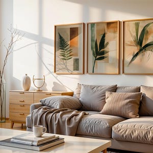 以下が含まれることがあります： 明るいグレーのコーナーソファ、抽象的な形をした植物のプリントが飾られたフレームが3つ、引き出し付きの木製のドレッサーがあるリビングルームのインテリア。 ソファには毛布とクッションが置かれています。 部屋はミニマルなスタイルで、自然光が差し込んでいます。