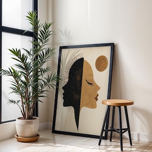 Puede incluir: Una impresión enmarcada en negro de una ilustración estilizada de dos caras, una negra y otra marrón, con un círculo dorado en el fondo. La impresión está colgada en una pared blanca junto a un taburete de madera con patas negras.