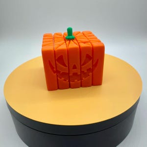 Könnte beinhalten: Eine orangefarbene, würfelförmige Halloween-Dekoration, die einem Kürbisgesicht nachempfunden ist. Der Würfel hat ein geschnitztes Gesicht und einen grünen Stiel. Die Dekoration steht auf einer gelben Oberfläche.