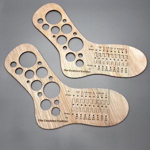 Op de afbeelding: Twee houten sokkenbreipatronen met gaten van verschillende grootte en meetmarkeringen. De tekst "The Enabled Knitter" is op elke meter gedrukt. De meters zijn lichtbruin en op een grijze achtergrond.