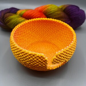 May include: An orange knitted bowl with a small opening on the side. A skein of colorful yarn in shades of purple, orange, green, and yellow is behind the bowl. The bowl is likely handmade.