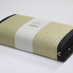 Angled top-down view of an Agura tatami cushion, designed for Zen-style cross-legged sitting. The surface uses natural igusa (rush grass) from Kumamoto, with soft green tones and black edging for a calm, refined design. A simple branded package from a 150-year-old tatami maker is wrapped at the center. A side handle allows for easy carrying. The firm cushion helps align the pelvis and provides comfort during extended meditation or mindfulness sessions.