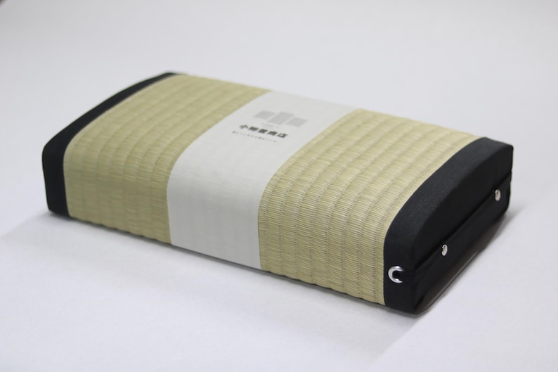 Angled top-down view of an Agura tatami cushion, designed for Zen-style cross-legged sitting. The surface uses natural igusa (rush grass) from Kumamoto, with soft green tones and black edging for a calm, refined design. A simple branded package from a 150-year-old tatami maker is wrapped at the center. A side handle allows for easy carrying. The firm cushion helps align the pelvis and provides comfort during extended meditation or mindfulness sessions.