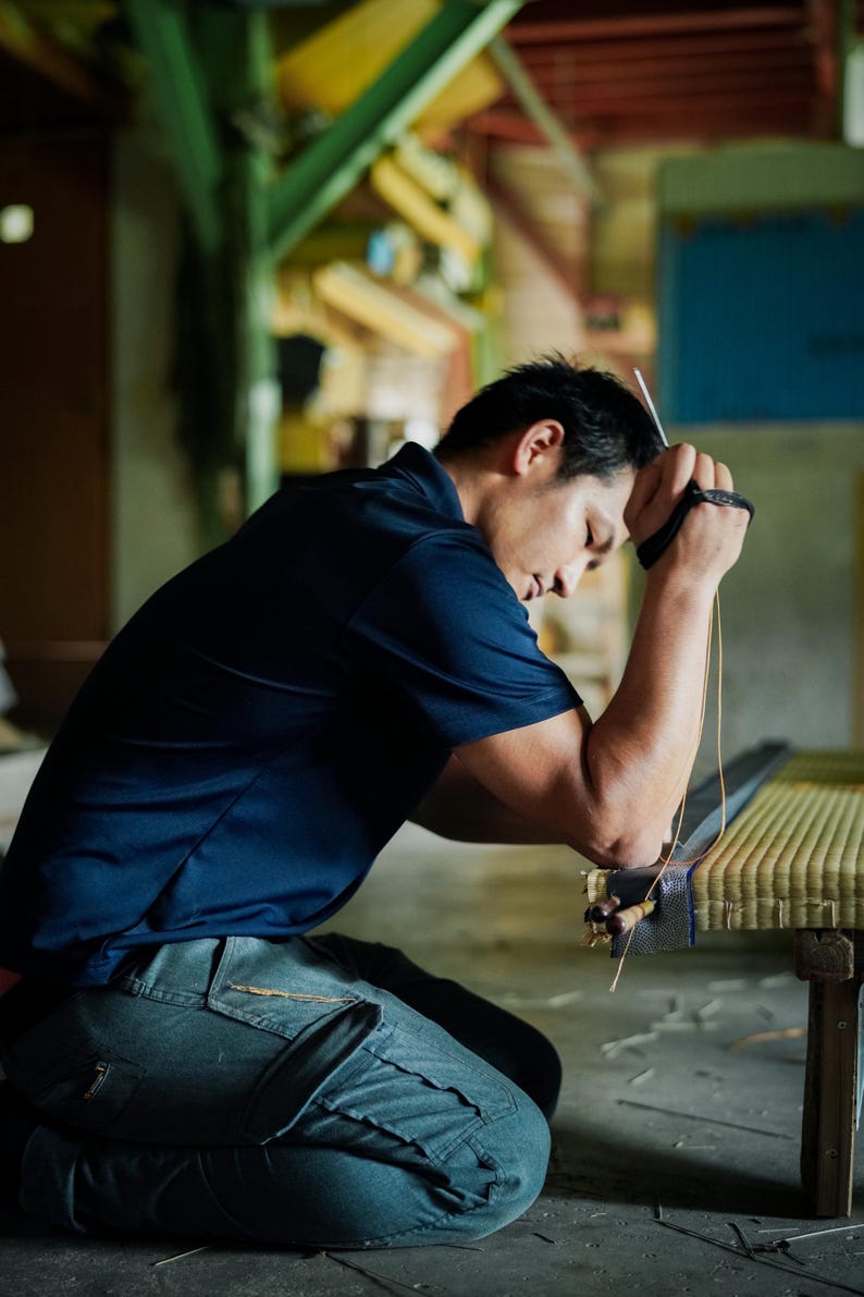 Ryuji Koyanagi, the fifth-generation craftsman of Koyanagi Tatami, a traditional tatami shop with over 150 years of history, is seen meticulously finishing a tatami mat in his workshop. With a sharp, focused gaze, he hand-stitches the mat’s edge, embodying the deep pride and sincere dedication that define Japanese craftsmanship.