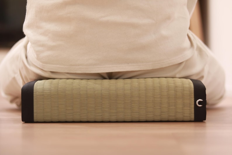 A rear-facing shot showing a model seated cross-legged (a traditional Japanese Zen posture) on a tatami cushion. The vivid green of natural igusa and the sleek black borders create a striking contrast. The model is firmly seated, and the ample thickness and moderate firmness of the cushion visibly stabilize the pelvis. Its wide and stable form makes it well-suited for long meditation or mindfulness sessions.