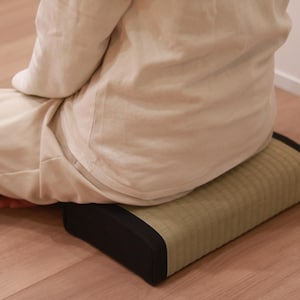 A shot taken from a diagonal rear angle showing a model sitting cross-legged (a traditional Japanese Zen posture) on a tatami cushion. The cushion features a refined design with the vivid green hue of natural igusa (rush grass) and contrasting black edges. With moderate firmness and stable support, the cushion helps align the posture and firmly support the pelvis. Its calm, grounded appearance is ideal for Zen and meditation practice.