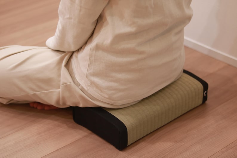 A shot taken from a diagonal rear angle showing a model sitting cross-legged (a traditional Japanese Zen posture) on a tatami cushion. The cushion features a refined design with the vivid green hue of natural igusa (rush grass) and contrasting black edges. With moderate firmness and stable support, the cushion helps align the posture and firmly support the pelvis. Its calm, grounded appearance is ideal for Zen and meditation practice.