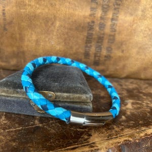 Puede incluir: Una pulsera de cuero trenzado azul con un cierre plateado. La pulsera presenta hebras de cuero azul claro y oscuro. La pulsera se exhibe sobre una superficie de estilo vintage.
