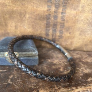Puede incluir: Pulsera de cuero trenzado en tonos marrón y negro, con cierre plateado. La pulsera es circular y parece sin usar. El fondo es un entorno de estilo vintage, con una superficie de cuero y una caja de terciopelo. Diámetro: aprox. 6 cm.