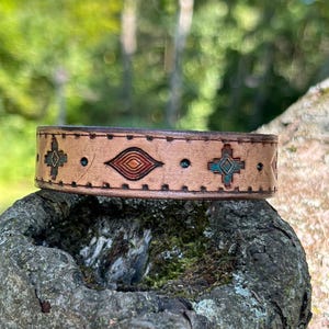 May include: A tan leather cuff bracelet featuring a repeating pattern of embossed geometric shapes, including diamonds and crosses, accented with small teal and orange painted details and a stitched border along the top and bottom edges.