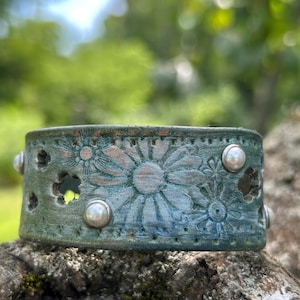 Puede incluir: Una pulsera de cuero envejecido de color turquesa con un diseño floral. La pulsera presenta margaritas en relieve y formas recortadas. Tachuelas plateadas están espaciadas uniformemente en los bordes. La pulsera descansa sobre una superficie musgosa y texturizada.