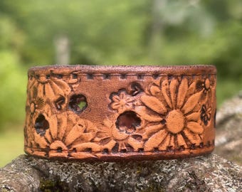 May include: A wide, tan-colored leather cuff bracelet featuring a deeply embossed floral pattern with large daisy-like flowers and small circular cutouts. The edges of the leather band are finished with a decorative stitched border detail.