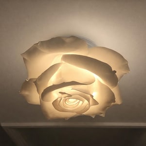 May include: A white, rose-shaped ceiling light fixture. The light is on, illuminating the petals and creating a warm glow. The fixture is mounted on a white ceiling, and the overall design is elegant and decorative.