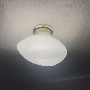 May include: A ceiling-mounted light fixture with a frosted glass shade in an oval shape. The fixture has a gold-colored metal base and is mounted on a textured white ceiling. The light is on, illuminating the room.