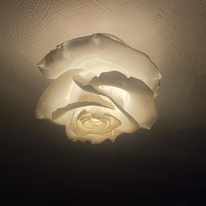 May include: A white, rose-shaped ceiling light fixture. The light is on, illuminating the delicate petals. The fixture is mounted on a textured ceiling, creating a soft, ambient glow. The design is elegant and floral.