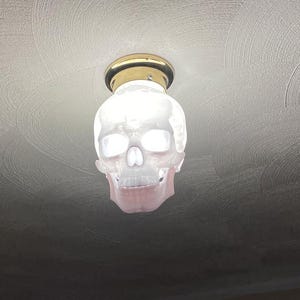 May include: A skull-shaped light fixture with a white, translucent shade and a gold-colored base. The fixture is mounted on a textured, off-white ceiling. The skull's eye sockets and nasal cavity are visible.