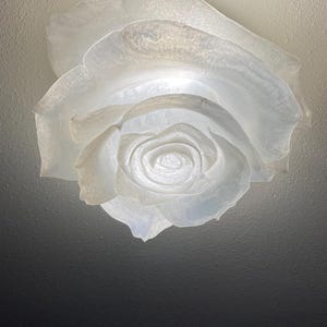May include: A white ceiling light fixture shaped like a blooming rose. The translucent material creates a soft, diffused glow from within, highlighting the layered, curved petals that form the flower structure against a plain ceiling surface.