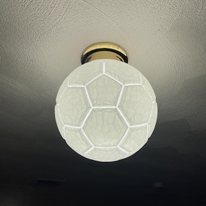 May include: A white, soccer ball-shaped ceiling light fixture. The light has a gold-colored base and is mounted on a textured white ceiling. The light is illuminated, and the room is dark.