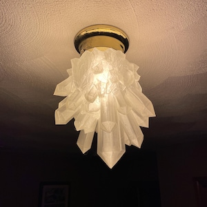 May include: A ceiling light fixture featuring a cluster of translucent, white, crystal-shaped glass shards arranged in a downward-pointing formation. The fixture is attached to the ceiling with a circular, polished gold-tone metal base.
