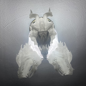 May include: A white, 3D-printed light fixture shaped like a stylized animal skull. The fixture has sharp teeth and pointed horns, with a bright light emanating from within, creating a dramatic effect. The fixture is mounted on a ceiling.
