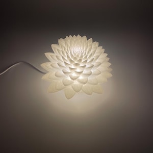 May include: A white, illuminated flower-shaped lamp. The lamp has multiple layers of petals, creating a textured, layered appearance. The light emanates from the center, casting a soft glow. A white cord extends from the lamp.