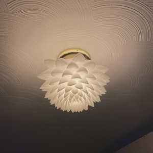 May include: A white, flower-shaped ceiling light fixture with layered petals. The light has a gold-colored base and is mounted on a textured white ceiling. The room is dimly lit.