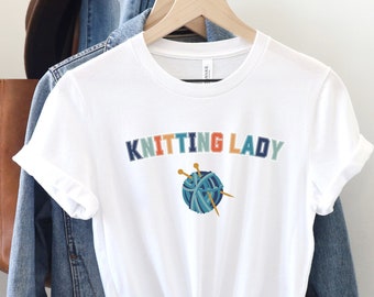 May include: White t-shirt with the text "Knitting Lady" in colorful letters and a graphic of a ball of yarn with knitting needles.