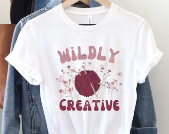 May include: White t-shirt with a pink and red graphic design that says "WILDLY CREATIVE" with a ball of yarn and knitting needles.
