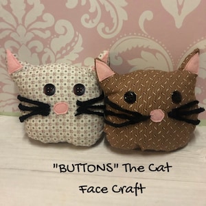 May include: Sew by number learn to sew kit for kids featuring two cat face craft projects. The kit includes two fabric cat faces, one brown and one white, with black yarn for whiskers and pink felt for noses. The kit is titled "BUTTONS" The Cat Face Craft.