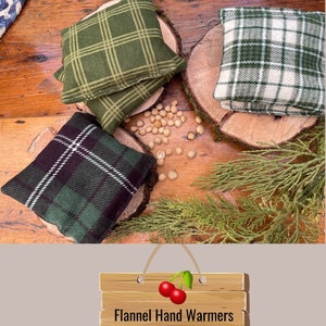 May include: Four flannel hand warmers in green plaid patterns, made from organic cherry pits. The warmers are displayed on wooden slices with sprigs of greenery and scattered cherry pits. A wooden sign with the text "Flannel Hand Warmers made from Organic Cherry Pits" hangs below.