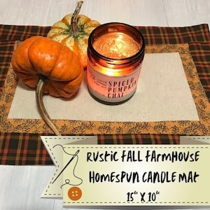 May include: A rustic fall farmhouse homespun candle mat in brown and orange plaid with a cream center. The mat is decorated with a small orange pumpkin and a candle with a label that reads "Spiced Pumpkin Chai". The mat measures 15 inches by 10 inches.