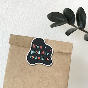 May include: A black sticker with a white outline in the shape of a cloud with the text "it's a good day to knit" in colorful letters and small red hearts.