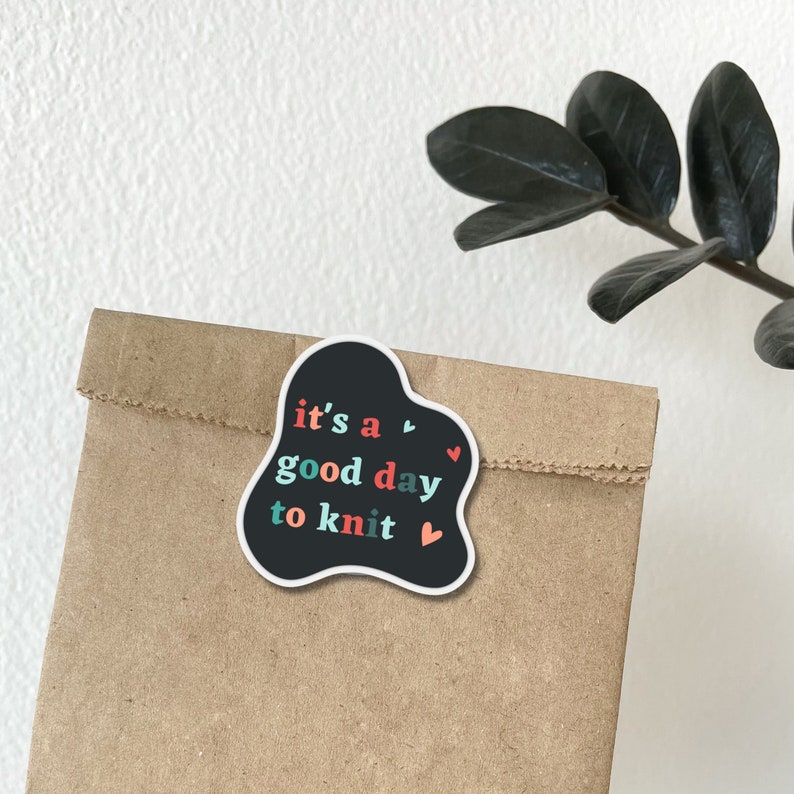 May include: A black sticker with a white outline in the shape of a cloud with the text "it's a good day to knit" in colorful letters and small red hearts.