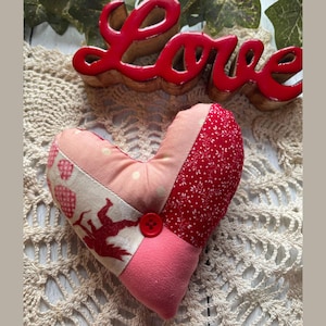 Puede incluir: Una almohada con forma de corazón hecha de retazos con un botón rojo en el centro. La almohada está hecha de tela rosa, roja y blanca con diferentes patrones. La almohada está sobre un encaje blanco de ganchillo.