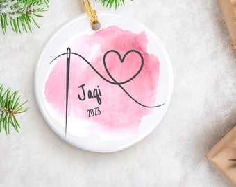 May include: White ceramic Christmas ornament with a pink watercolor background. A black needle and thread design forms a heart shape. The name "Jagi" and the year "2023" are printed in black. A gold string is attached for hanging.