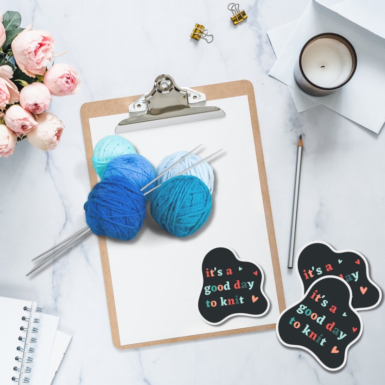 May include: A flat lay image of a clipboard with blue yarn balls and knitting needles. There are two stickers on the table that say "it's a good day to knit" with a heart symbol.