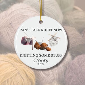 May include: White ceramic Christmas ornament with a knitting theme. It says "Can't talk right now, knitting some stuff Cindy 2024". The ornament features a drawing of knitting needles, yarn, and a bowl of yarn.