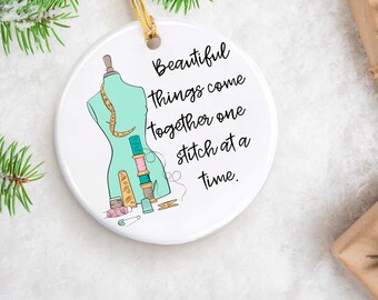 May include: White ceramic ornament with a green sewing dummy and colorful spools of thread. The ornament has the text "Beautiful things come together one stitch at a time."