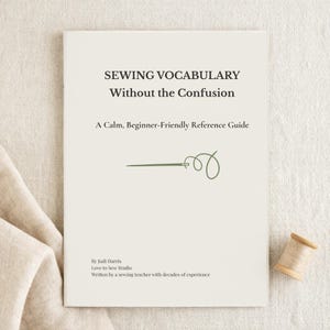 May include: A cream-colored book titled "SEWING VOCABULARY Without the Confusion" with a green needle and thread illustration. The book is a beginner-friendly reference guide. A wooden spool of thread sits nearby.