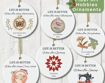 May include: Six round white ceramic ornaments with gold string hangers. Each ornament features a different fiber art hobby with a watercolor illustration and text that reads "Life is better when I'm doing [hobby]" with a name and the year 2024. The hobbies featured are embroidery, needlepoint, scrapbooking, quilting, cross-stitch, and sewing.
