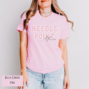 May include: Pink Bella Canvas t-shirt with the text "NEEDLE POINT Maria" in white and pink lettering.