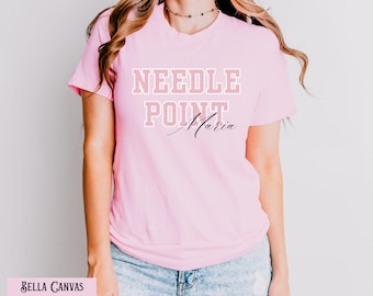 May include: Pink Bella Canvas t-shirt with the text "NEEDLE POINT Maria" in white and pink lettering.