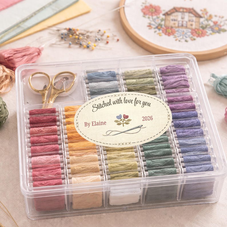 Puede incluir: Una caja de pl&aacute;stico transparente llena de hilo de bordar colorido, unas tijeras doradas y una etiqueta que dice "Stitched with love for you By Elaine 2026". El hilo est&aacute; dispuesto en filas de varios tonos.