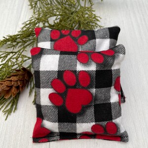 May include: Two black and white buffalo plaid fabric heat packs with red paw prints. The heat packs are stacked on top of each other.