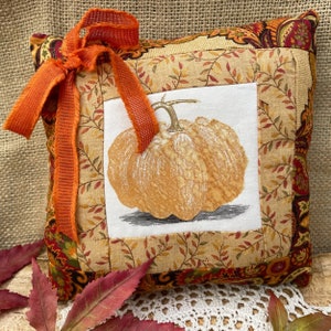 May include: A decorative pillow with a patchwork design featuring a brown and white pumpkin illustration. The pillow has a brown, orange, and red fabric with a floral pattern. A bright orange ribbon is tied around the pillow.