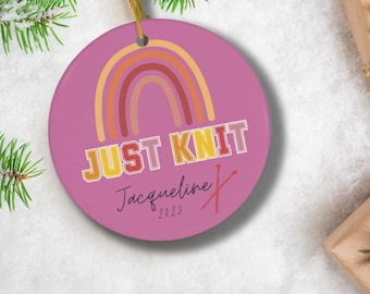 May include: A pink round Christmas ornament with a rainbow design and the text "JUST KNIT" in yellow, red, and orange letters. The name "Jacqueline" and the year "2023" are written in black cursive script below the text. Two red knitting needles are crossed below the name.
