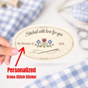 Op de afbeelding: Ovale sticker met kruissteekontwerp. De sticker heeft de tekst "Stitched with love for you" en "By Momma El 2026". Het heeft een kruissteek bloemenontwerp met een naald en draad. De woorden "Personalized Cross-Stitch Sticker" zijn ook zichtbaar.