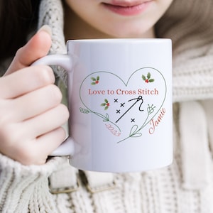 May include: White ceramic mug with a green heart design featuring a cross stitch needle and the text "Love to Cross Stitch" and "Jamie" with the year "2023" below the heart.